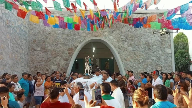 A crowd of local people celebrating a traditional religious festival in Rio Lagartos, carrying a saint's statue in front of a stone church with colorful banners overhead.