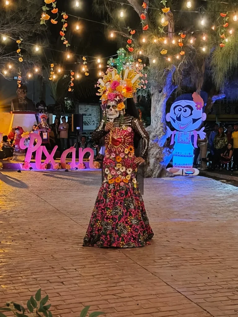 Una persona luciendo un elaborado y monumental disfraz de Catrina, con un vestido floral de corte sirena y un tocado de flores con una cruz, posando en el parque de Río Lagartos durante el concurso de disfraces del Festival de Ánimas.