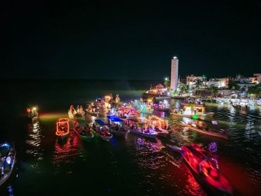 Vista aérea de la procesión náutica nocturna del Festival de Ánimas, con docenas de lanchas iluminadas en la ría de Río Lagartos y el faro del puerto al fondo.