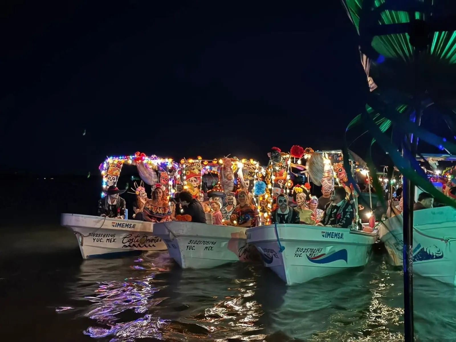 Tres lanchas decoradas con luces y altares del Día de Muertos flotando en la ría de Río Lagartos durante el Paseo de Pixanes nocturno.