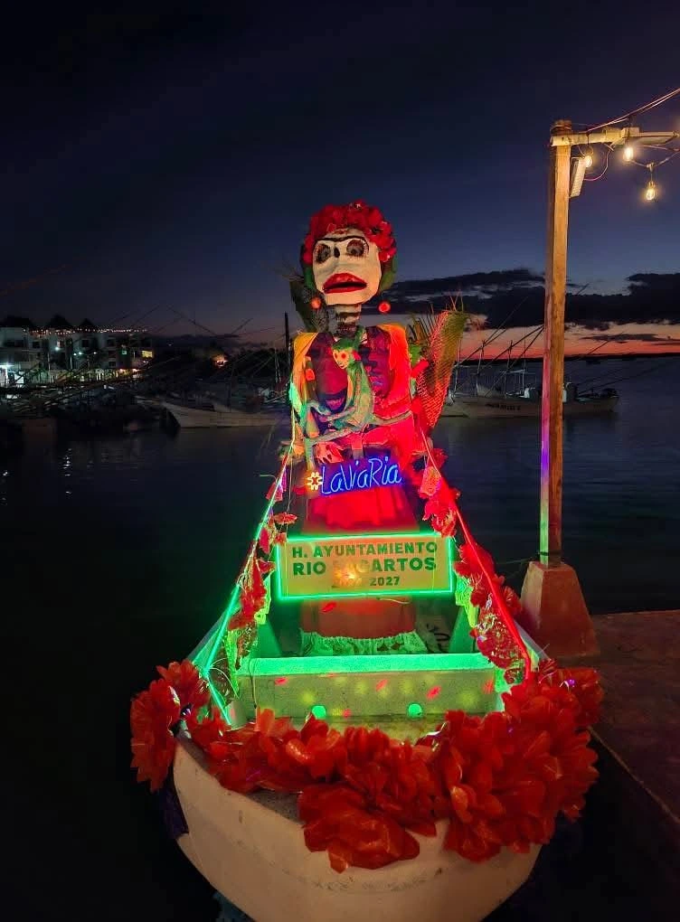 Decorated tour boat featuring a large traditional Catrina figure illuminated by neon lights during the Festival of Ánimas in Río Lagartos at sunset.