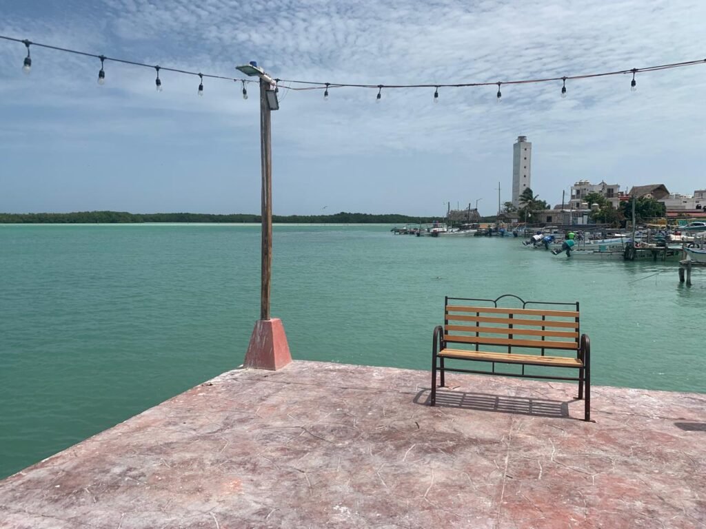 A peaceful stone pier in Rio Lagartos with a wooden bench, decorative lights, and a view of the lighthouse across the turquoise lagoon.