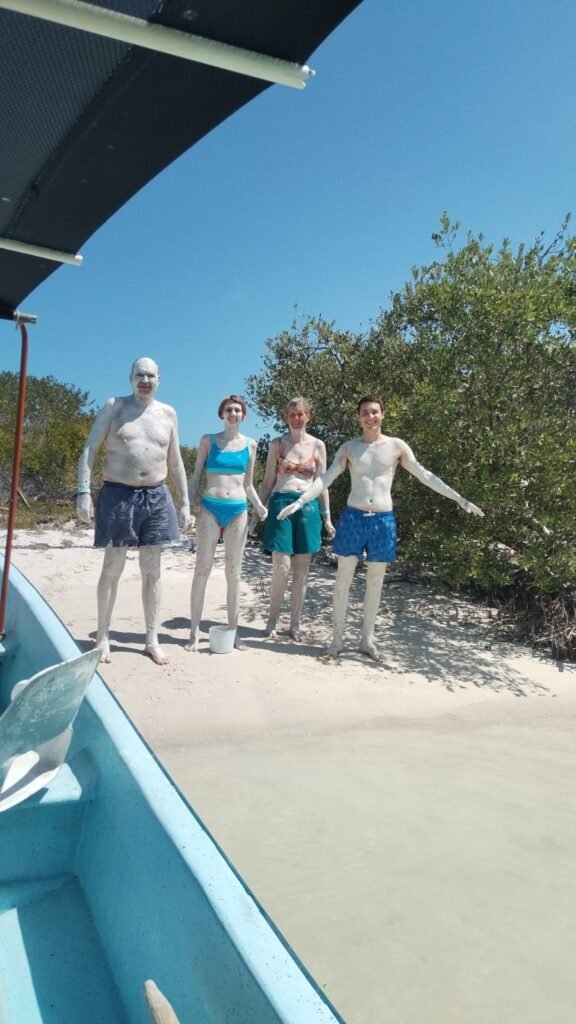 Four tour guests covered in white medicinal mud pose smiling on a beach next to a boat in Rio Lagartos, Yucatan, after enjoying the Mayan Bath Rio Lagartos ritual.